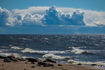 Beautiful seascape. Cloudy sky. Reflection of sunlight on sea waves. Stones on sandy beach.