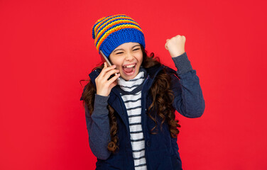 glad kid talking on phone in hat on red background. teen girl in down vest with smartphone.