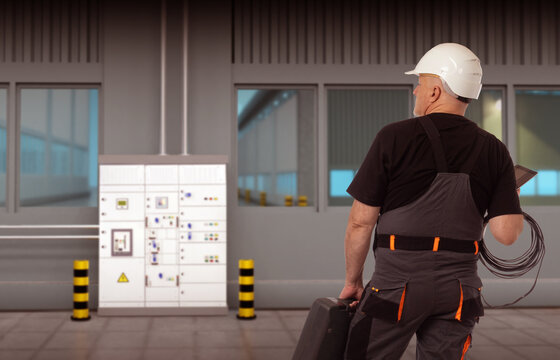 Electrician in production room. Man go to electrical panel. Factory worker with wires stands in hangar. Electrician man with his back to camera inside building. Electrification of industrial buildings
