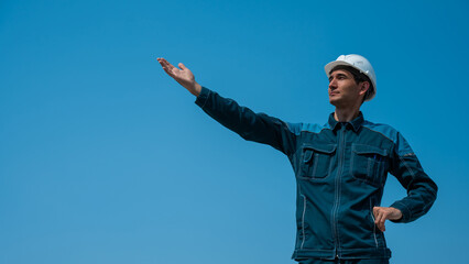 A caucasian man in work clothes and a construction helmet stands against the background of a blue sky with his arms outstretched to the sides.