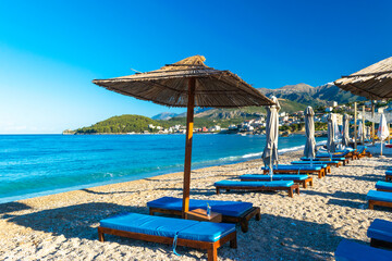 Landscape of beautiful clean sand and pebble beach with beach umbrellas and sun loungers on mountains background. Himare. Albania.