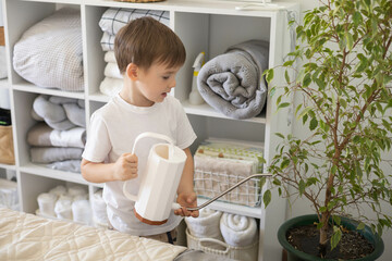 Little cute boy is watering indoor plants from a stylish watering can in a designer home interior. © kostikovanata