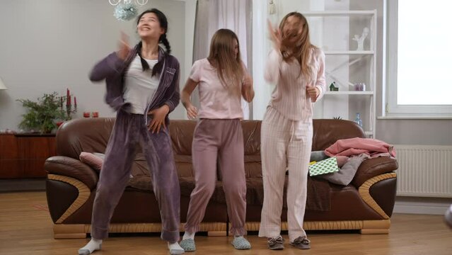 Wide shot three joyful women dancing in living room on pajama party singing smiling. Portrait of carefree relaxed Asian and Caucasian friends having fun enjoying weekend leisure indoors