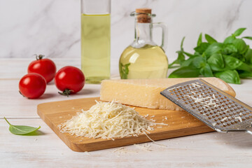 Heap of grated parmesan on a cutting board. Grana padano cheese whole and grated, hand grater, olive oil, tomatoes and green basil herb on a white wooden table. Delicious hard cheese.