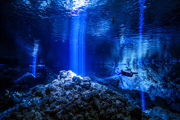 A cave diver descends into a cave system with light rays penetrating the water in the background.