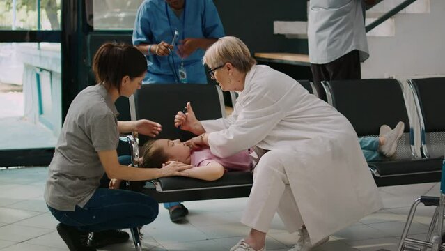 Senior medic consulting little unconscious girl with flashlight, doing checkup in health center lobby. Doctor trying to help young kid with illness, fainting on chairs in hospital reception.