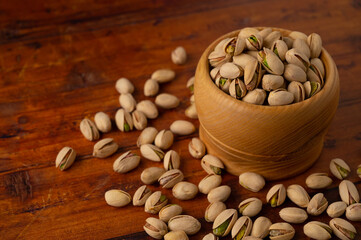Roasted and salted pistachios in a wooden bowl surrounded by scattered pistachios on a brown wooden table background.