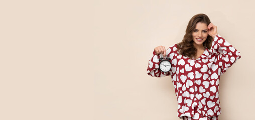Attractive brunette in red and white heart pajamas with an alarm clock in her hand on a light brown...