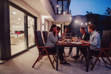 Fototapeta na wymiar A group of young diverse people having dinner on the terrace of a modern house in the evening. Fun for friends and family. Celebration of holidays, weddings with barbecue.