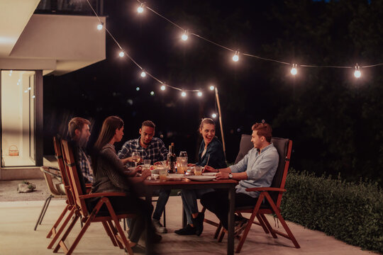 A Group Of Young Diverse People Having Dinner On The Terrace Of A Modern House In The Evening. Fun For Friends And Family. Celebration Of Holidays, Weddings With Barbecue.