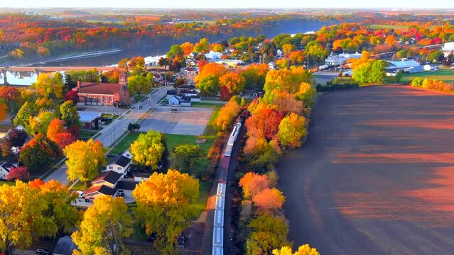 Freight train rolls through small town, all ablaze with Fall colors, aerial view.
