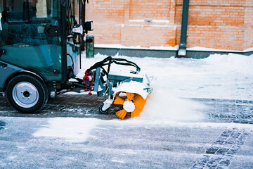 City service cleaning snow, a small tractor with a rotating brush clears a road in the city park from the freshly fallen snow on a winter day. Service. Vehicle. Cleanup. Snowfall. Season. Shoveling