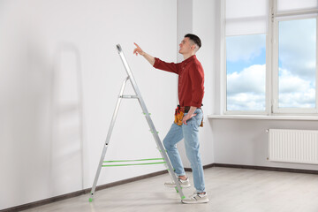 Young handyman with stepladder and tool belt working in room