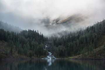 Mountain creek flows from forest hills into glacial lake. Tranquil scenery with rocks in clearance of mysterious fog. Small river and coniferous trees reflected in calm alpine lake in early morning.