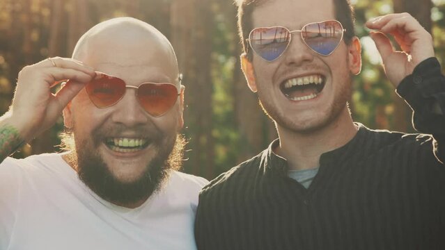 Homosexual couple smile nad laugh. Portrait of two mixed race gay men in love wearing sunglasses of heart shape smiling to camera. LGBTQI, Pride Event, LGBT Pride Month, Gay Pride Symbol.