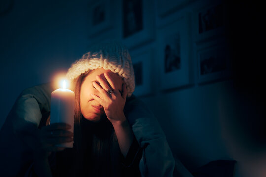 Unhappy Woman Feeling Cold Sitting In The Dark Using A Candle. Person Freezing In Her Apartment Warming In A Blanket

