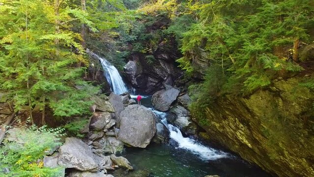 Aerial along river in rocky gorge of lush green forest with waterfall and lone hiker observing falls from boulder
