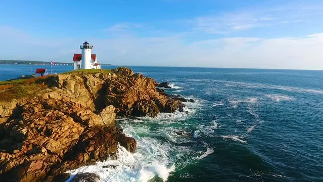 Aerial towards Waves crash over rocks aerial over Maine island with lighthouse and view of homes on mainland