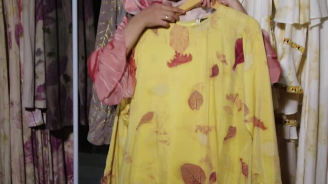 an Asian Muslim woman, showing her Ecoprint craft, in the form of a yellow long dress. Standing among the neatly arranged dresses.
