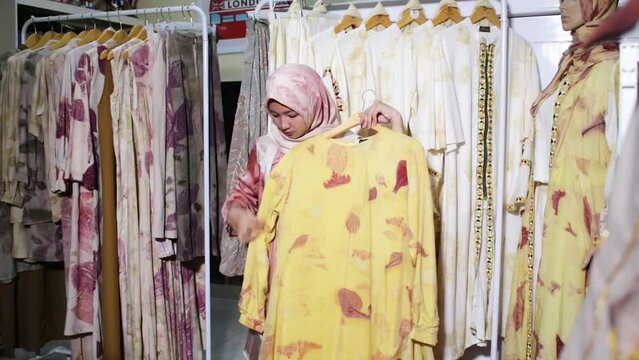an Asian Muslim woman, showing her Ecoprint craft, in the form of a yellow long dress. Standing among the neatly arranged dresses.