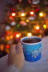 A woman's hands in a sweater hold a blue cup of coffee by the window on the background of Christmas tree. Christmas lights. Hot winter flavored drink. - 553626499