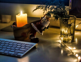 desktop with computer decorated with Christmas decor candles, garland, santa claus in the evening dark background - 553626652