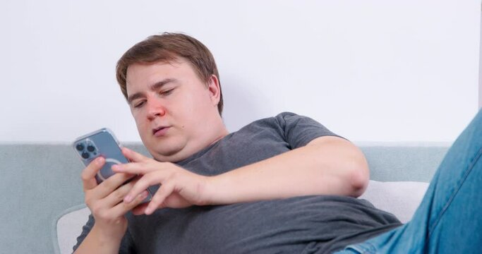 Adult plump man lies on couch in room reading news on smartphone. Brown-haired guy with red runny nose enjoys resting in room sticking to phone