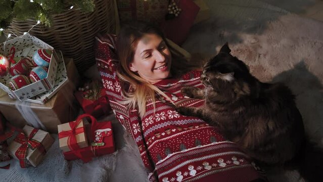 The atmosphere of the new year. A young woman in the company of her favorite cat is lying under a Christmas tree. Concept of Christmas celebration. Slow-motion