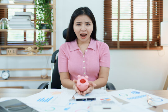 Portrait of an Asian female businesswoman showing a serious and anxious expression in an SME business , but no money in the pink pig piggy bank , saving plans