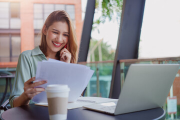 Young businesswoman working with papers and talking on mobile phone, reading financial documents while sitting at cafe.