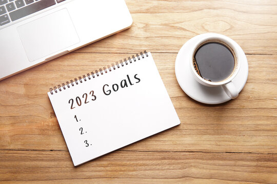 2023 goal list, motivational words in notebook on table with laptop and cup of coffee.