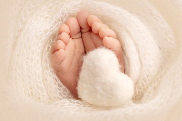 The tiny foot of a newborn baby. Soft feet of a new born in a white wool blanket. Close up of toes, heels and feet of a newborn. Knitted white heart in the legs of a baby. Macro photography. 