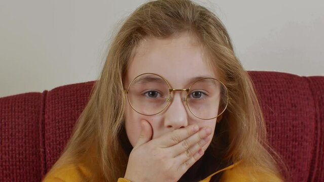 Funny pretty girl with long blond hair wearing round big glasses and yellow sweatshirt. Girl seriously looks at camera then smiles showing a toothless mouth and funny covers her mouth with her palm