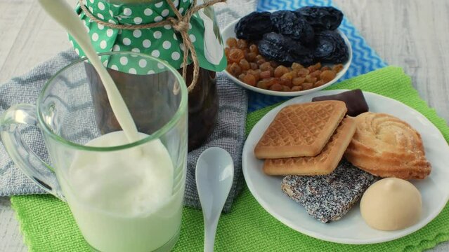 Fresh natural milk is poured into a glass. Nearby on multi-colored napkins is a jar of jam, a white teaspoon, a saucer with dried fruits and a saucer with cookies and sweets. Dairy food concept