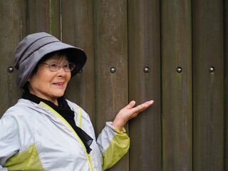 木製の壁を背景に手で案内する高齢日本人女性