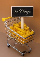 WORLD HUNGER text on Blackboard label against Shopping trolley cart Filled With Pasta on Beige background. Food and groceries shopping price increase, Rising food cost food crisis inflation concept. 