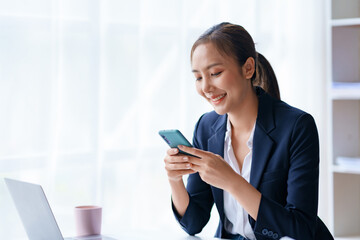 Beautiful Asian businesswoman is using smartphone to chat with customers at work and play various social media applications in a relaxed smile and good mood.