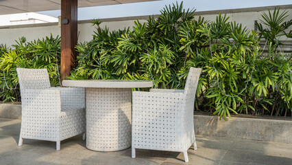 A place to rest. Two white wicker chairs and a table stand on the paved terrace. Nearby are lush ornamental green plants.