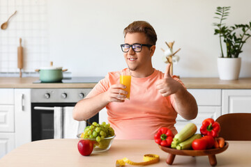 Young overweight man with glass of juice showing thumb-up in kitchen