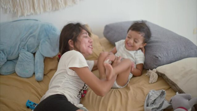 Brunette latin mother tickling her son smiling and laughing after waking up on a bed with beige sheets on a warm morning