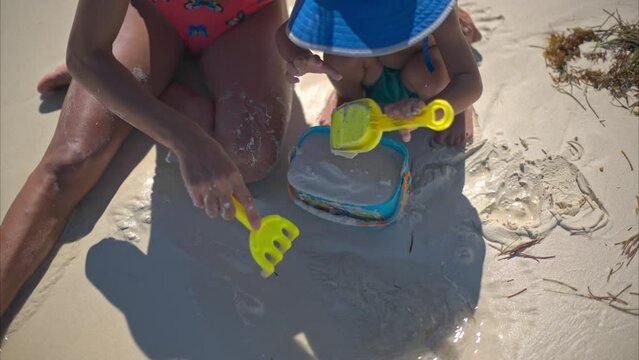 Cute young baby boy playing with his mother on the sand at the beach shoveling sand into a bucket