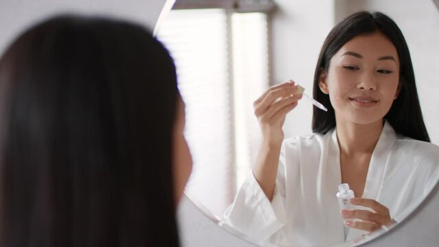 Young asian lady applying nourishing serum on face, smiling to her mirror reflection at home in morning, free space