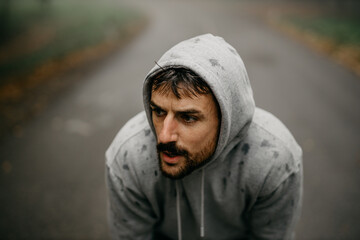 Fototapeta premium Young Caucasian athletic man pulling the hood over his head while having sports training in nature during the cold autumn day.