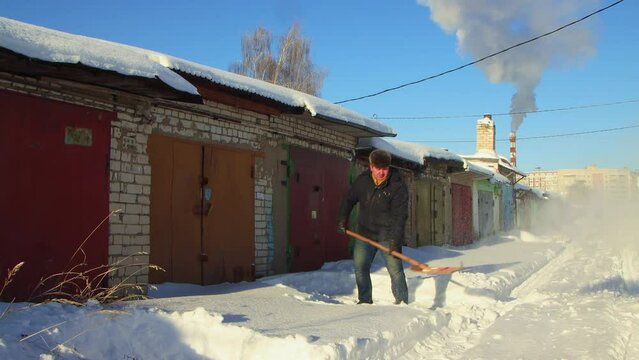 A man deftly clears the passage to the garage from snow with a wooden shovel. Large snowdrifts around after a snowstorm, sunny day. City life, heavy snowfall. UHD 4K.