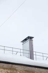 Smoke, rectangular, metal exhaust pipe on the roof of a modern house. The roof of the building is covered with white snow against the gray sky. Cloudy, cold winter day, soft light.