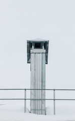 Smoke, rectangular exhaust pipe on the roof of a modern house. The roof of the building is covered with white snow against the gray sky. Cloudy, cold winter day, soft light.