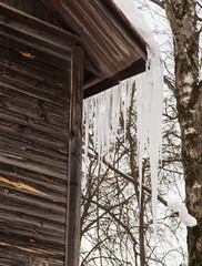 Sharp, frozen icicles hang on the corner of the roof, winter or spring. Log wall of an old wooden house with windows. Large cascades of icicles in smooth, beautiful rows. Cloudy winter day, soft light