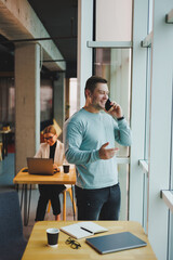 Business man, manager looking out of window in deep thought, standing at workplace with phone in office, thinking about future of company, thinking about solution for work task