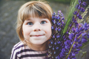 Cute adorable little toddler girl with huge bouquet of blossoming violet lupnies flowers. Portrait of smiling preschool child in domestic garden on warm spring or summer day. Summertime.