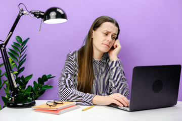 Portrait of tired frustrated secretary employee young woman in casual shirt sitting work at white office desk using laptop internet prop up face, posing over purple color background wall in studio